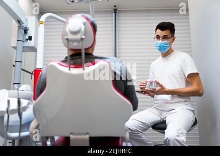 Le chirurgien dentaire avec la couronne dentaire dans les mains modèle parle au patient au sujet des prothèses. Homme au rendez-vous du dentiste Banque D'Images