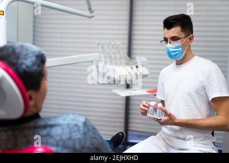 Le chirurgien dentaire avec la couronne dentaire dans les mains modèle parle au patient au sujet des prothèses. Homme au rendez-vous du dentiste Banque D'Images
