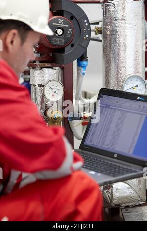 Thème de l'industrie de l'énergie : un technicien en combinaison rouge et casque blanc vérifie les paramètres de chauffage sur l'ordinateur portable. Entretien et inspection. Banque D'Images