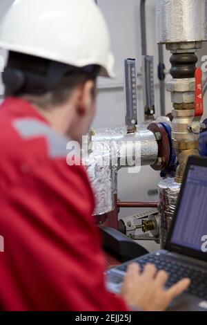 Thème de l'industrie de l'énergie : un technicien en combinaison rouge et casque blanc vérifie les paramètres de chauffage sur l'ordinateur portable. Entretien et inspection. Banque D'Images