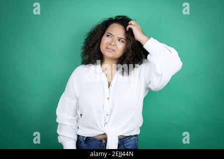 Une jeune fille afro-américaine portant une chemise blanche sur fond vert trouble et se demande s'il y a des questions. Incertain avec le doute, pensant avec la main sur h Banque D'Images