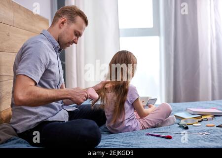 le père fait une coupe de cheveux pour sa fille en utilisant le peigne à la maison sur le lit, beau homme caucasien assis s'occupant de son enfant fille. à l'intérieur Banque D'Images