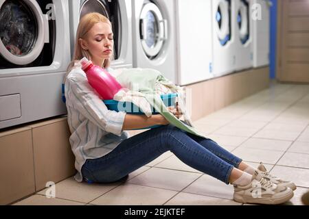 Femme ennuyée tenant des vêtements sales et bouteille rose de détergent dans le lavabo, assis sur le sol incliné sur la machine à laver, assis enfoncé seul, dans le ho de lavage Banque D'Images