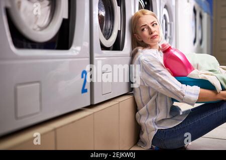 Femme fatiguée faisant la lessive, s'assoit sur le sol, n'a aucune force pour rien, femme se pencha sur la machine à laver en attente, après une dure journée de travail Banque D'Images