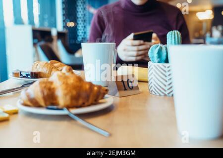 Une tasse de thé chaud à la vapeur et des croissants frais sur la table où un jeune homme utilise son téléphone pendant son petit déjeuner au restaurant de l'hôtel. Mise au point sélective, Banque D'Images