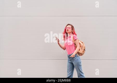 Jeune femme blogueuse aux poils teints roses restant près du mur gris et utilisant un smartphone Banque D'Images