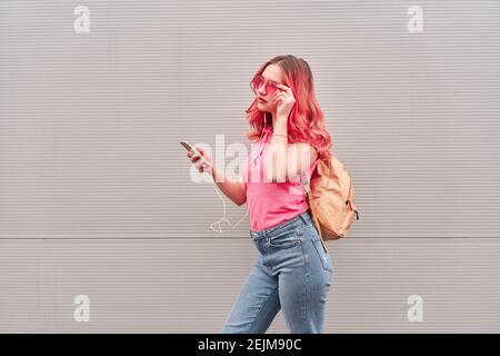 Jeune femme blogueuse aux poils teints roses restant près du mur gris et utilisant un smartphone Banque D'Images