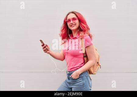 Jeune femme blogueuse aux poils teints roses restant près du mur gris et utilisant un smartphone Banque D'Images