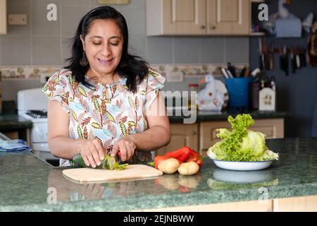 Femme hispanique d'âge moyen souriante qui fait de la salade fraîche en elle cuisine - dame en cuisine saine avec des légumes biologiques Banque D'Images