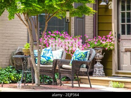 Coin salon extérieur confortable avec chaises en osier et oreillers lumineux devant des pansies roses en pot sous l'arbre près de la fenêtre et porte avec chat peering dehors Banque D'Images