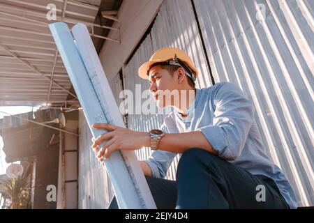 Homme d'architecture assis au sol et tenir le plan de conception des plans et porter un casque de sécurité lors de la construction du site par temps chaud. Architecture Banque D'Images