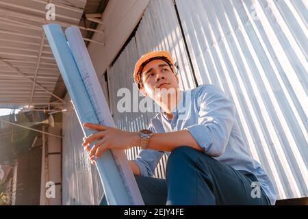 Homme d'architecture assis au sol et tenir le plan de conception des plans et porter un casque de sécurité lors de la construction du site par temps chaud. Architecture Banque D'Images