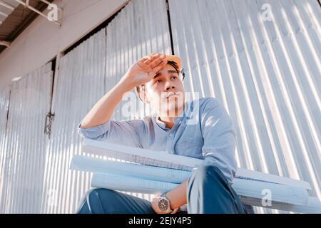 L'homme de l'architecture essuyait la sueur et lui assis sur le plan de conception de plans de mise au sol et de maintien et port d'un casque de sécurité à la construction du site dans chaud nous Banque D'Images