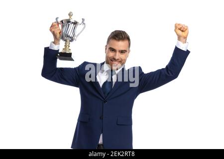 portrait d'un homme enthousiaste en costume bleu marine tenant les bras et trophée d'argent dans les airs célébrant la victoire et le sourire sur fond blanc en studio Banque D'Images