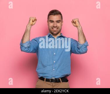 joyeux homme décontracté dans une veste en denim tenant les poings et célébrant la victoire, souriant et debout sur fond rose en studio Banque D'Images