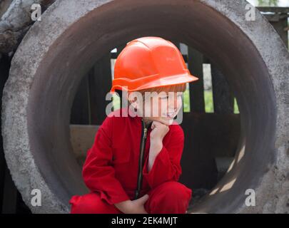 un garçon de 4 ans, mignon et curieux, dans un casque de construction orange et une combinaison de travail rouge, se trouve près de cercles de béton sur un chantier de construction. rires avec moquisme. Banque D'Images
