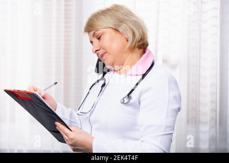 Le médecin caucasien Handsome femme avec stéthoscope parlant sur téléphone mobile, smartphone appels à la clinique, la vérification des soins de santé de la cliente sur papier Banque D'Images
