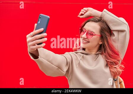 Jeune blogueuse avec des cheveux teints roses restant près du mur rouge et faisant selfie par smartphone Banque D'Images