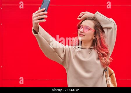 Jeune blogueuse avec des cheveux teints roses restant près du mur rouge et faisant selfie par smartphone Banque D'Images