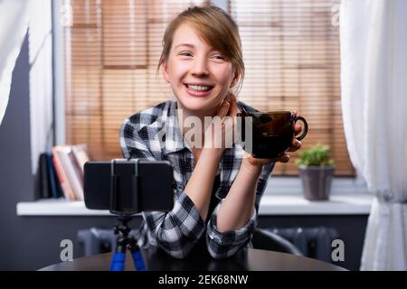 Femme blogueuse à côté d'un smartphone. La fille tient une tasse de thé dans ses mains. Elle enregistre la vidéo sur son gadget. Blogger femme diffuse la vidéo. En ligne Banque D'Images