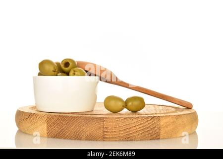 Plusieurs olives vertes piquantes bio savoureuses et épicées se trouvent dans une soucoupe en céramique blanche avec une cuillère en bois, sur un plateau rond en bambou. Le fond est blanc. Banque D'Images