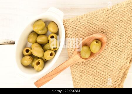 Quelques olives vertes piquantes bio savoureuses et épicées se trouvent sur une soucoupe en céramique blanche, une cuillère en bois et une serviette en jute sur fond de table blanche et naturelle. Banque D'Images