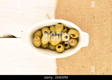 Quelques olives vertes piquantes bio épicées se trouvent sur une soucoupe en céramique blanche, une serviette en jute sur un fond de bois peint blanc naturel. Banque D'Images