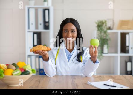 Faire des choix alimentaires sains. Belle nutritionniste féminine tenant croissant et pomme à table dans le bureau médical Banque D'Images