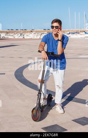 Un jeune homme reste avec un scooter électrique près de la mer dans la marina et parler sur un smartphone Banque D'Images