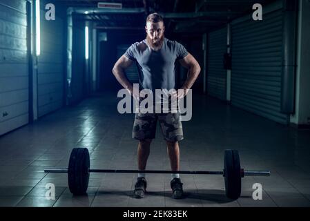 Image d'un bodybuilder sportif robuste se préparant à lui-même pour le levage lourd. Debout devant un poids lourd dans une pièce sombre. Banque D'Images