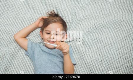 Petit garçon allongé sur le lit tenant un thermomètre dans la main et souriant. La température est normale. Soins de santé. Pandémie de coronavirus. Covid-19 Banque D'Images