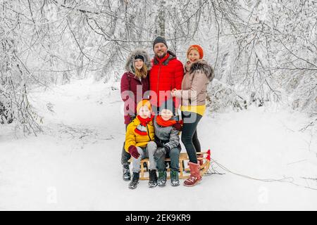 Parents souriants debout près d'enfants assis traîneaux dans la forêt pendant hiver Banque D'Images