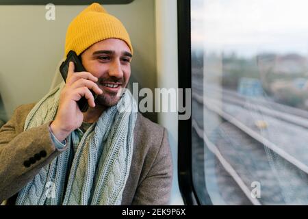 Homme souriant portant un chapeau en tricot parlant sur un téléphone portable pendant assis dans le train Banque D'Images