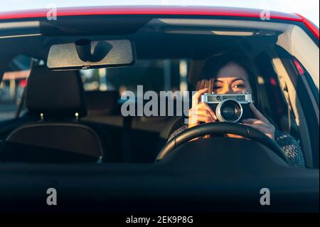 Jeune femme avec une caméra d'époque en voiture vue à travers le pare-brise par beau temps Banque D'Images