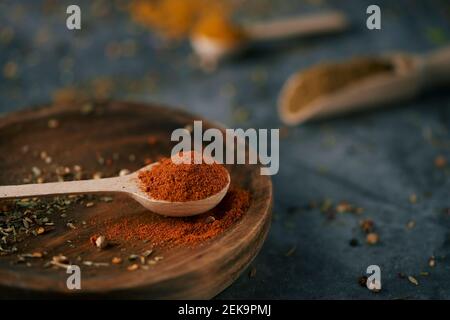 gros plan d'une cuillère en bois pleine de poudre de curry rouge, sur une assiette en bois, placée sur une surface en pierre sombre et quelques autres cuillères et pelles en bois avec une pâte à café Banque D'Images