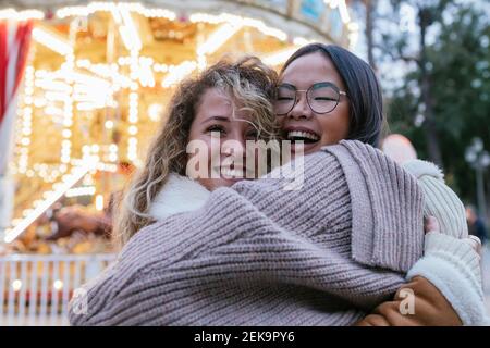 Rire des amies féminines s'embrassant contre un carrousel illuminé Banque D'Images