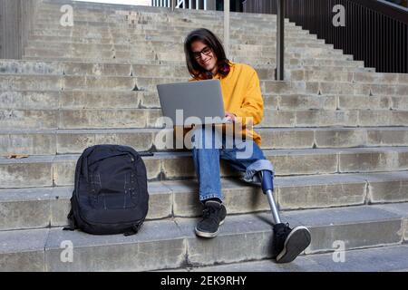 Homme souriant avec prothèse de jambe utilisant un ordinateur portable tout en étant assis sur étapes Banque D'Images