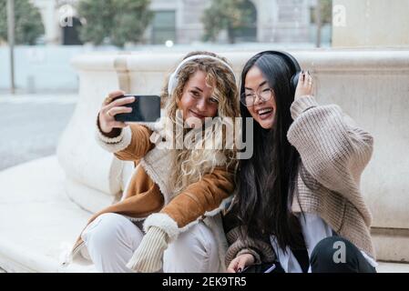 Des amies gaies emportant leur selfie sur leur smartphone tout en écoutant musique sur casque Banque D'Images