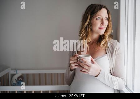 Femme enceinte attentionnée tenant une tasse de café tout en se tenant contre le berceau à la maison Banque D'Images