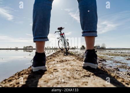 Vélo vu à travers les jambes d'une femme debout sur le sentier de randonnée au Delta de l'Ebro pendant la journée ensoleillée, Espagne Banque D'Images