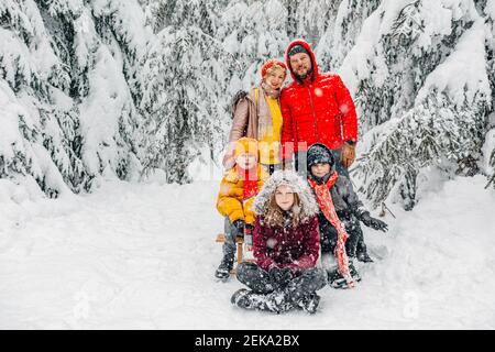 Parents debout par des enfants assis sur la neige dans la forêt Banque D'Images