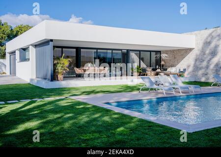 Chaises longues vides au bord de la piscine contre la maison moderne Banque D'Images