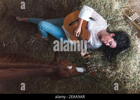 Femme adulte de taille moyenne avec un cheval jouant de la guitare tout en étant allongé foin en position stable Banque D'Images