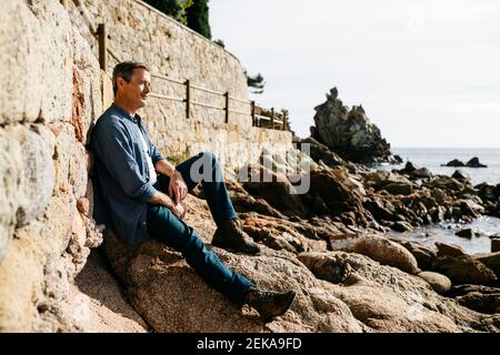 Homme attentionné assis sur le rocher à la plage pendant la journée ensoleillée Banque D'Images