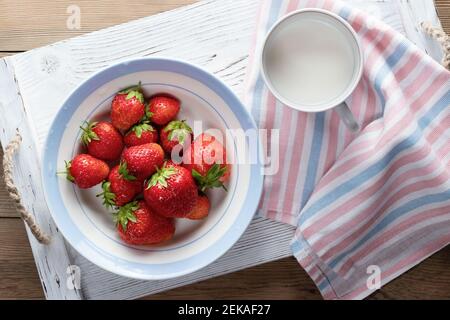 Fraises mûres naturelles dans un bol blanc et lait dans une tasse sur un plateau en bois blanc vue du dessus. Banque D'Images