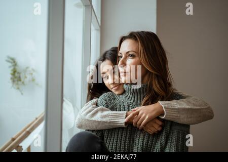 Fille embrassant la mère tout en contemplant par la fenêtre à la maison Banque D'Images