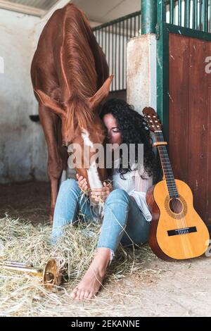 Femme à la guitare embrassant le cheval tout en nourrissant le foin en stable Banque D'Images