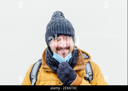 Homme souriant tirant le masque contre le mur blanc pendant COVID-19 Banque D'Images