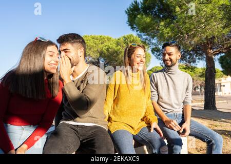 Deux jeunes couples hétérosexuels en train de se rire les uns les autres. Joyeux groupe millénaire de personnes qui s'amusent en plein air. L'homme parle à l'oreille de sa petite fille Banque D'Images