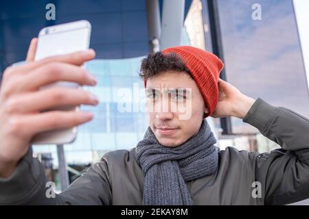 Jeune homme portant un chapeau tricoté qui prend le selfie à travers le téléphone mobile en ville Banque D'Images